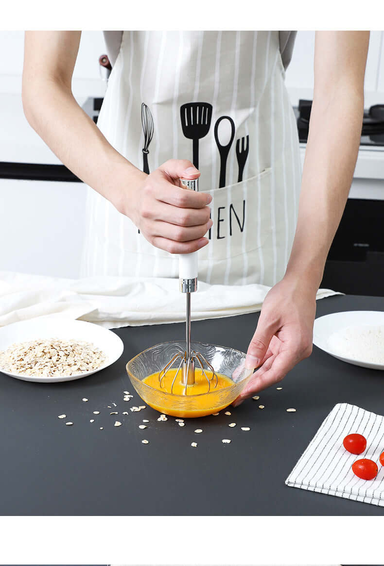 Hand whisking eggs in glass bowl with manual egg beater, kitchen accessories visible