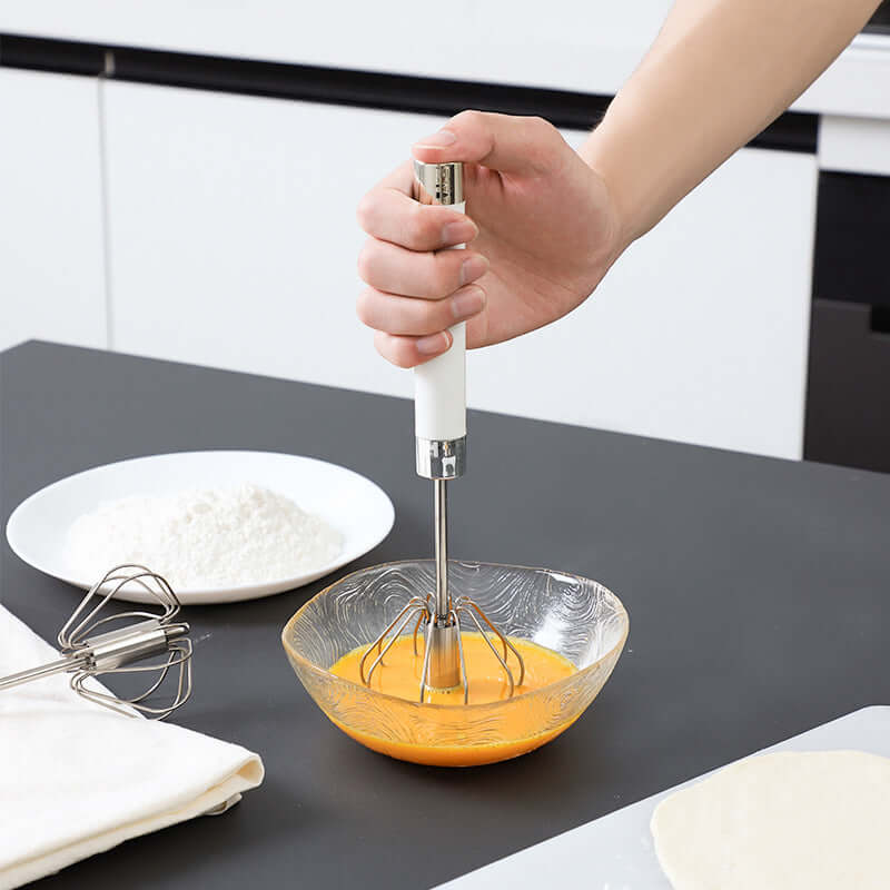 Hand using stainless steel egg beater whisk in glass bowl with eggs in modern kitchen