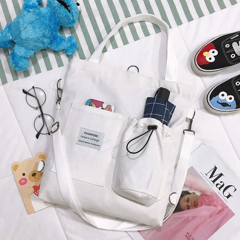 White canvas tote bag with outer pockets, glasses, plush toy, and painted sneakers on bed