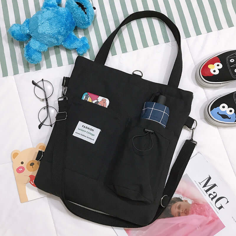Black canvas tote bag with pockets, glasses, plush toy, and playful shoes on bed