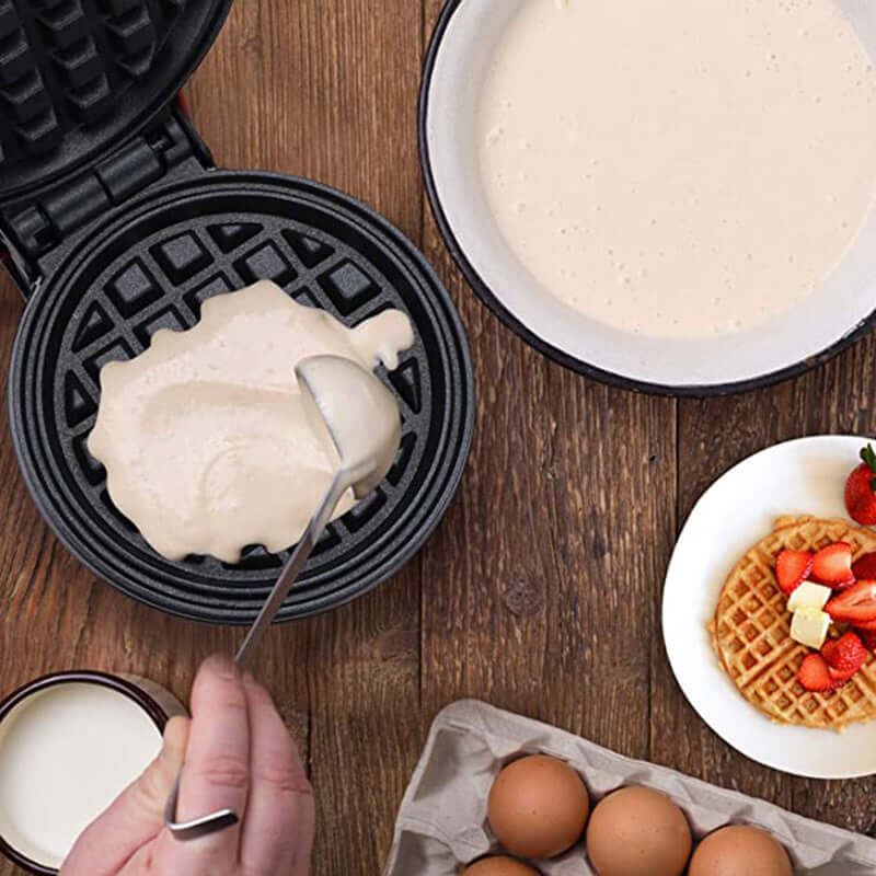 Waffle maker with batter being poured, eggs, milk, and plate of waffles with strawberries