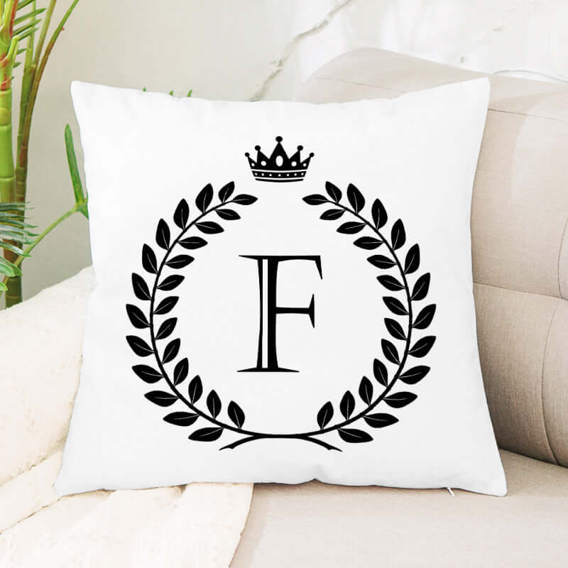 White decorative pillow with black letter F, laurel wreath, and crown design on a sofa.