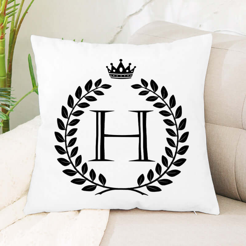 White decorative pillow with black H initial, laurel wreath, and crown design on a sofa