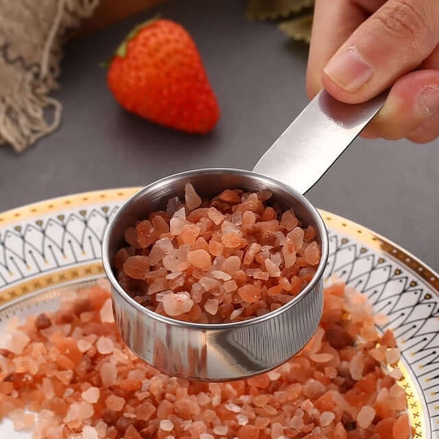 Hand holding measuring cup with pink Himalayan salt over a decorative plate in kitchen setting