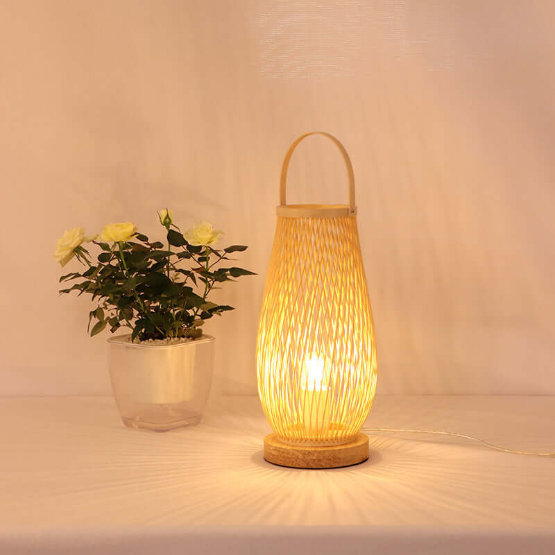 Wooden woven table lamp glowing beside potted white roses on a white surface