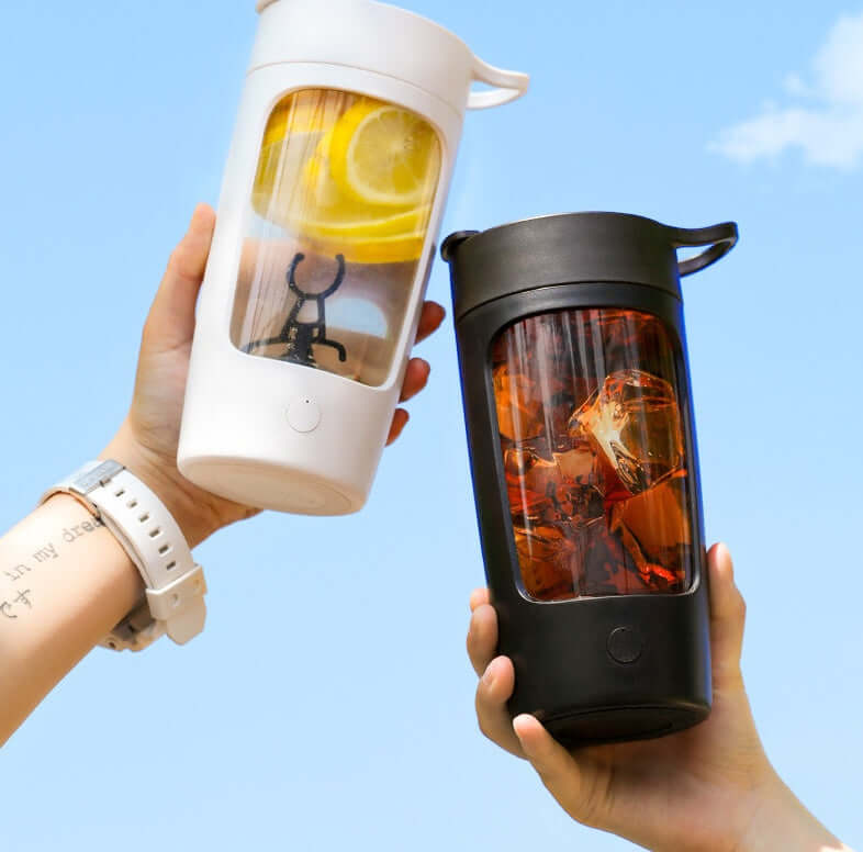 Two hands holding portable blender bottles, one with lemon slices, one with iced tea, against blue sky.