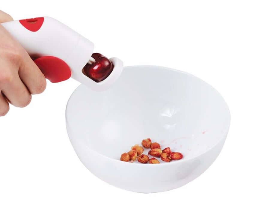 Hand using cherry pitter kitchen tool over white bowl with cherry pits