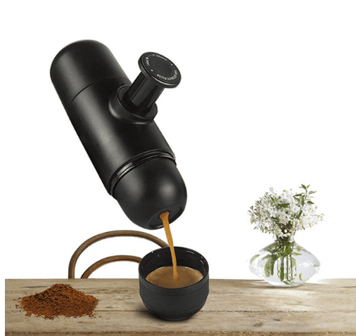 Manual portable coffee maker brewing espresso into a cup on a kitchen counter with ground coffee and a flower vase nearby.