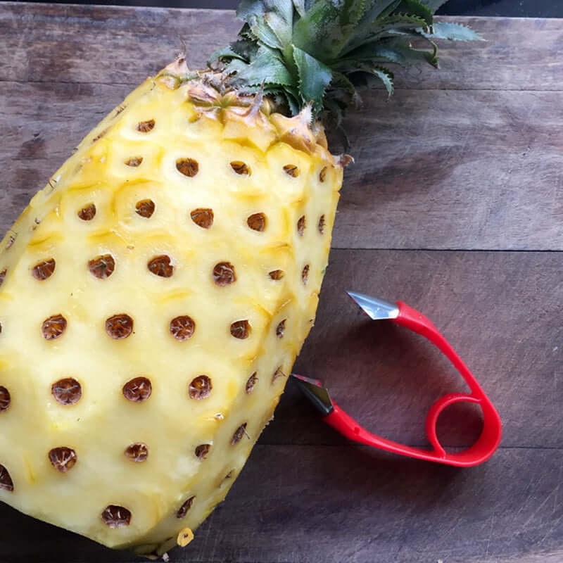 Pineapple eye remover tool with red handle beside peeled pineapple on wooden surface