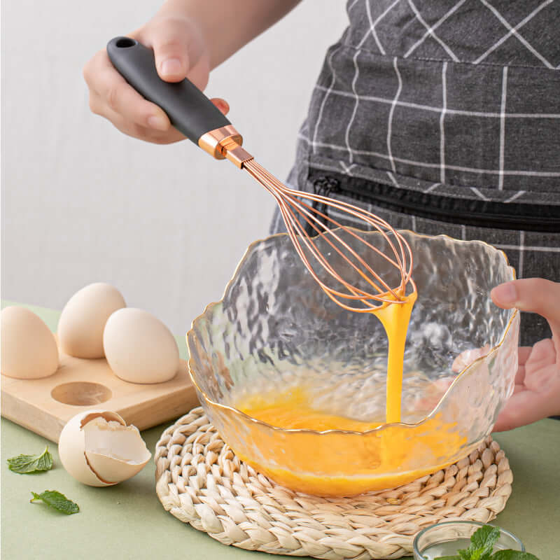 Hand whisking eggs in clear glass mixing bowl with kitchen whisk utensil on table