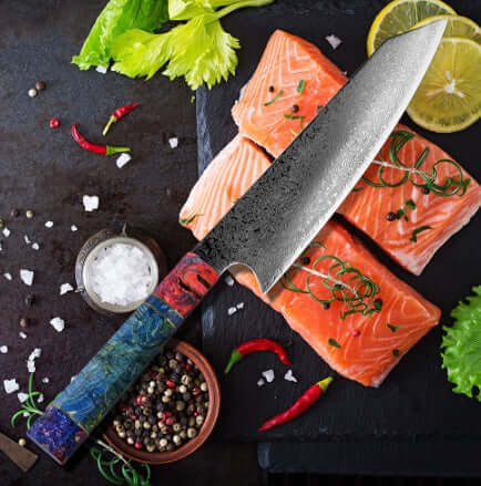 Damascus chef knife with colorful handle on raw salmon fillets, spices and lemon on black slate