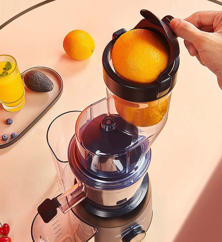 Hand placing orange in juicer machine with juice, avocado, and blueberries nearby