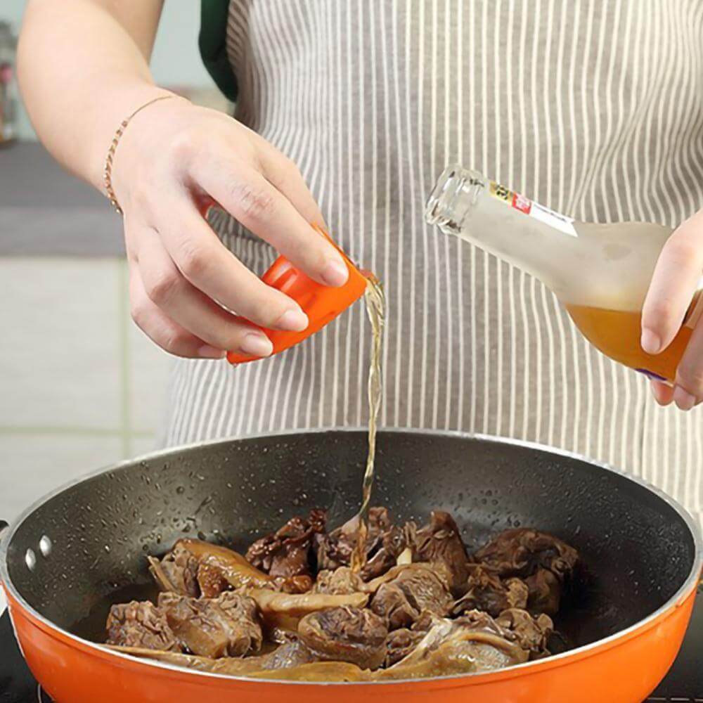 Person pouring sauce from bottle into red frying pan with meat, kitchen utensil in use