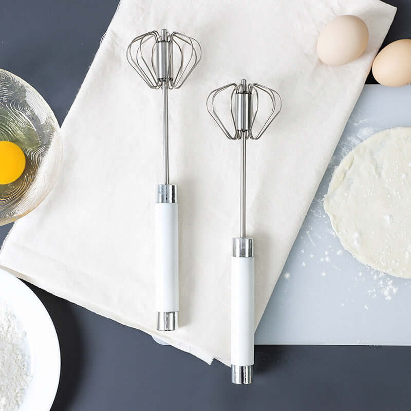 Stainless steel hand egg beaters with white handles on kitchen towel, surrounded by dough, flour, and eggs.