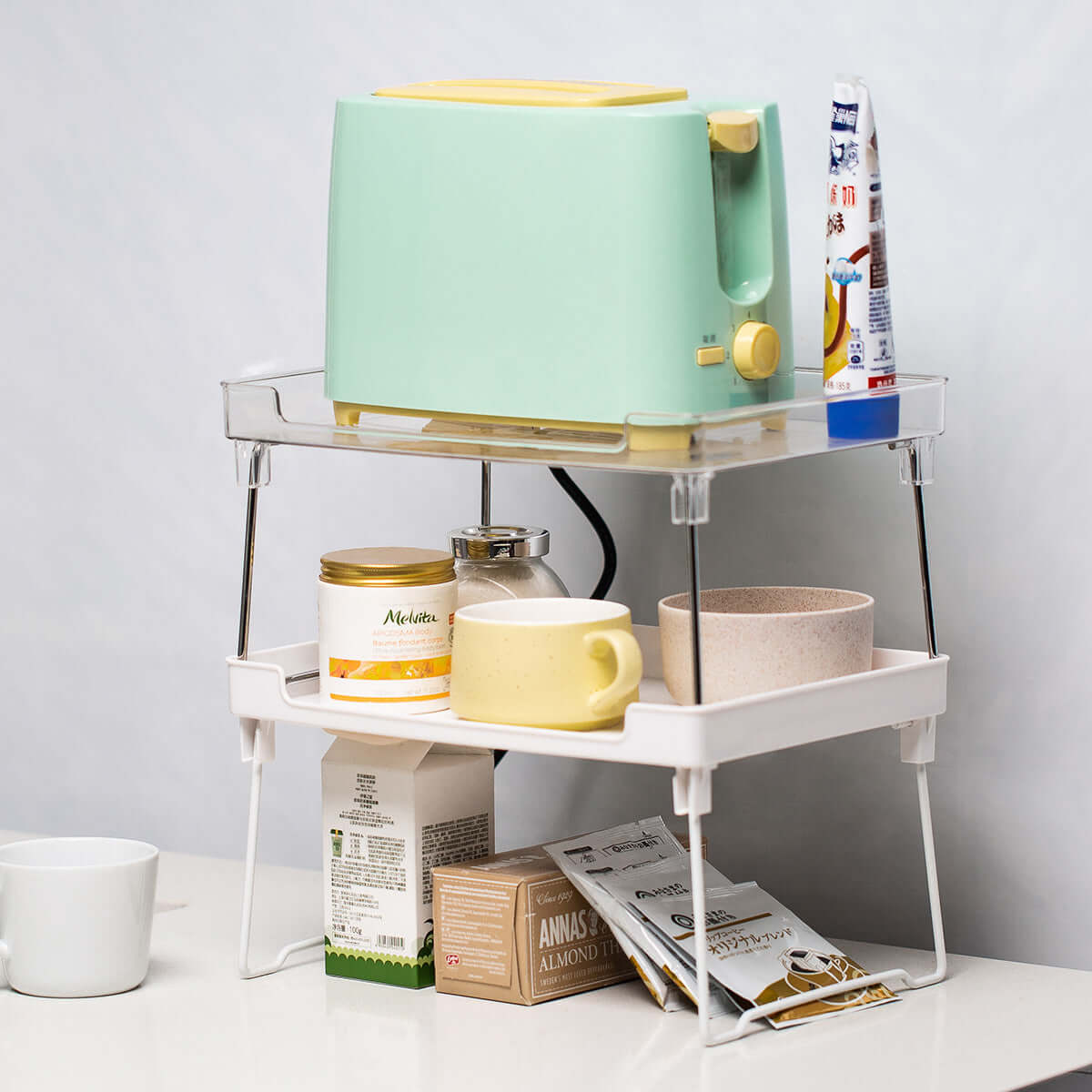 Two-tier kitchen organizer holding a green toaster, mugs, bowls, and pantry items on a counter