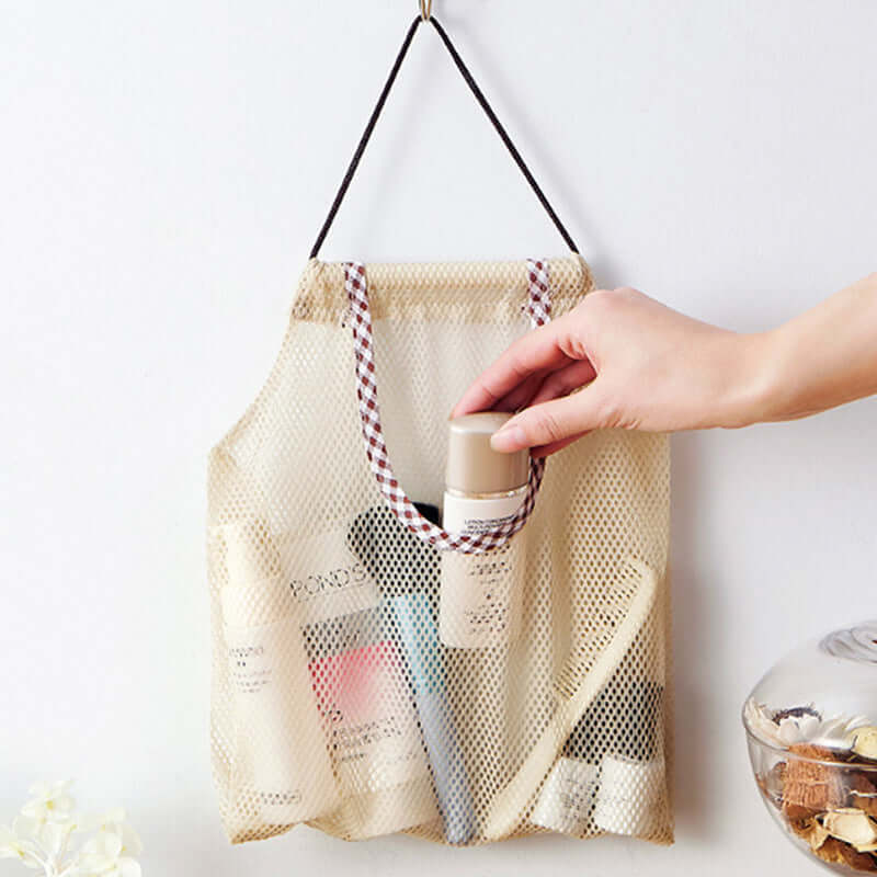 Hand placing bottle in hanging mesh storage bag with toiletries against a white wall