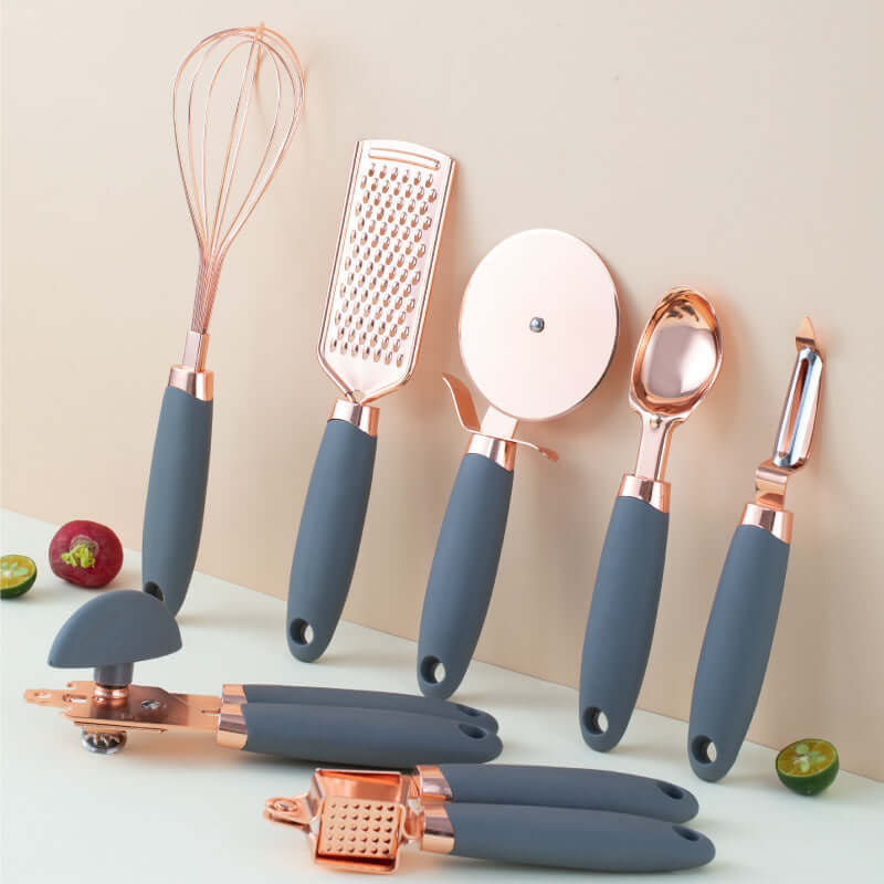 Set of rose gold kitchen utensils with gray handles, including whisk, grater, pizza cutter, can opener, garlic press, ice cream scoop, and peeler, displayed on a light surface.