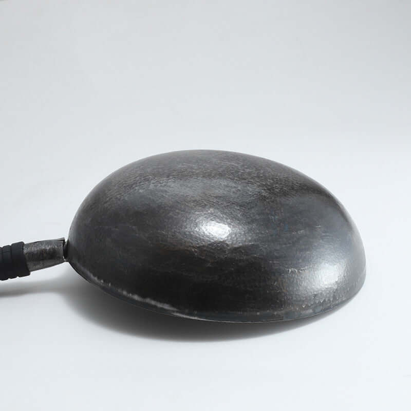 Hand-forged carbon steel wok with round dome shape and black handle on white background