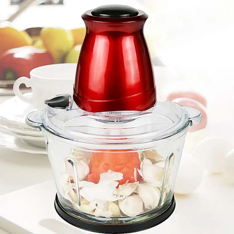 Electric food chopper with red top, glass bowl, garlic and tomato inside, on kitchen counter