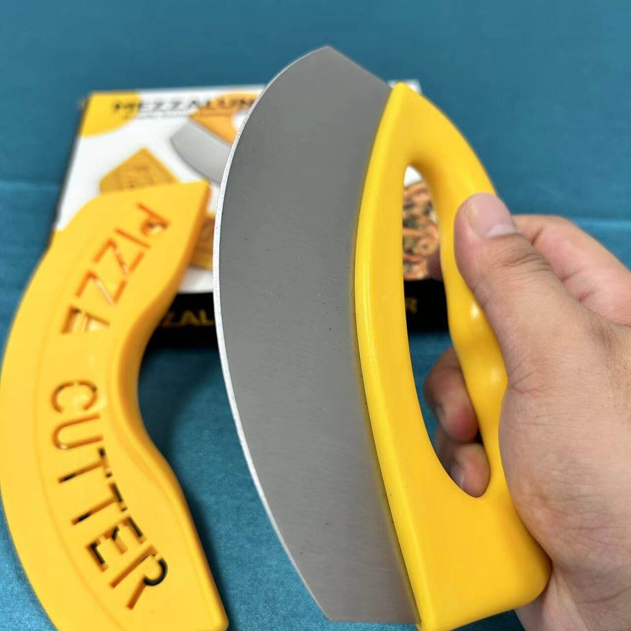 Hand holding a yellow mezzaluna pizza cutter with stainless steel blade on a blue surface