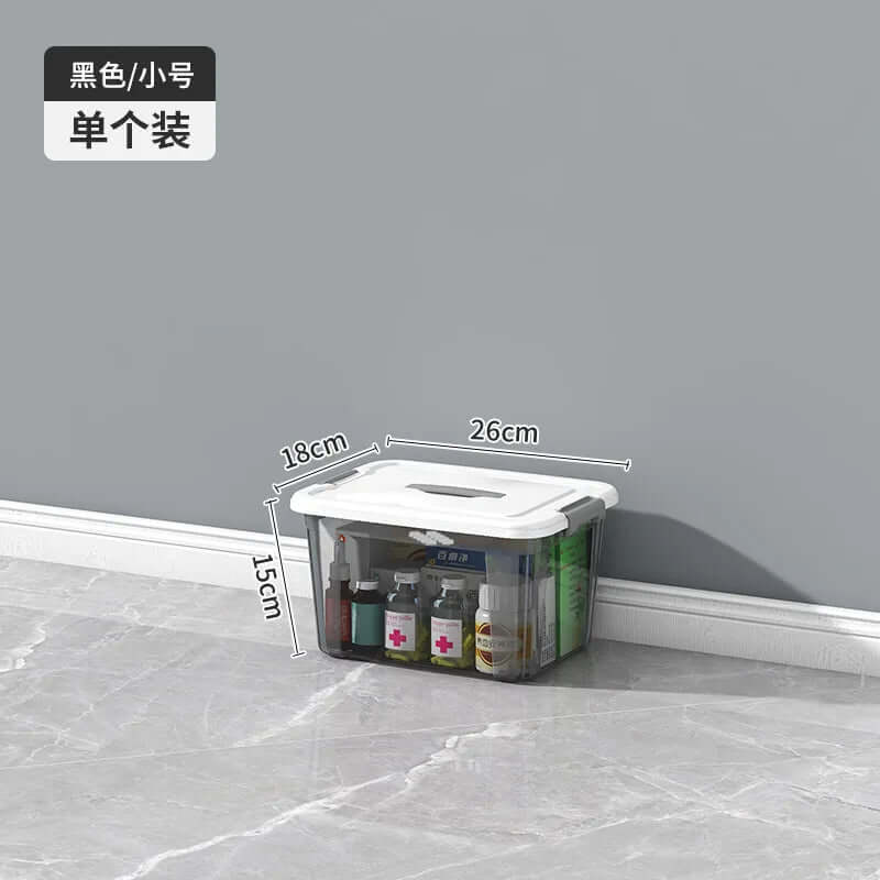 Clear plastic storage box with white lid, organizing medicine bottles, labeled with dimensions 26cm x 18cm x 15cm, on tiled floor against a gray wall.