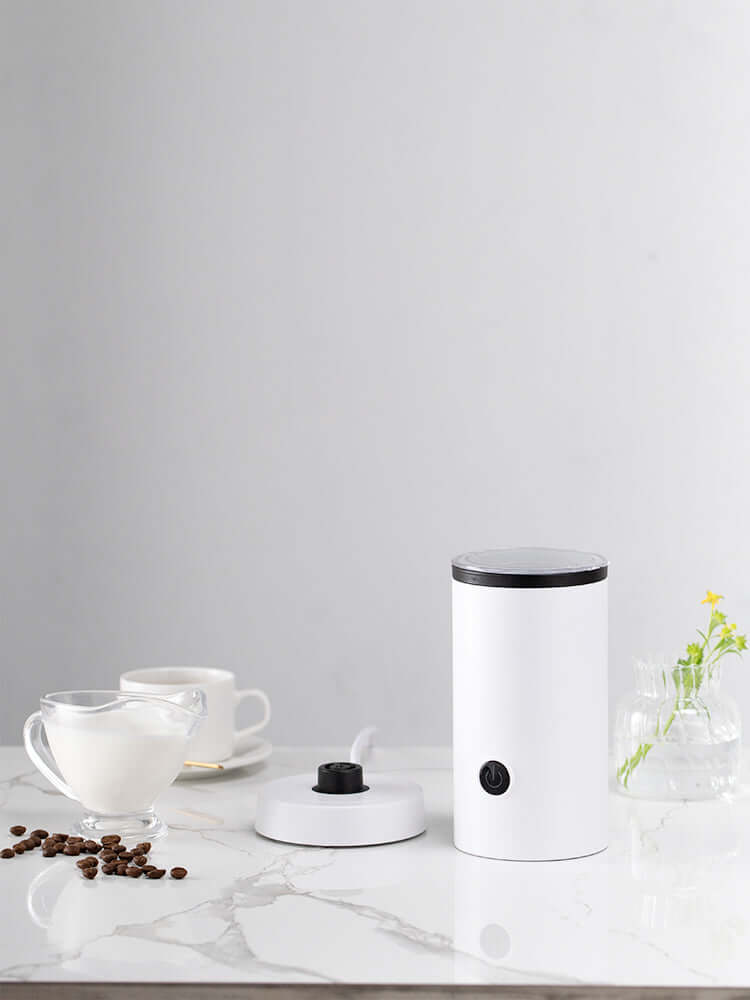 Electric milk frother on kitchen counter with coffee beans, cup, and glass milk jug