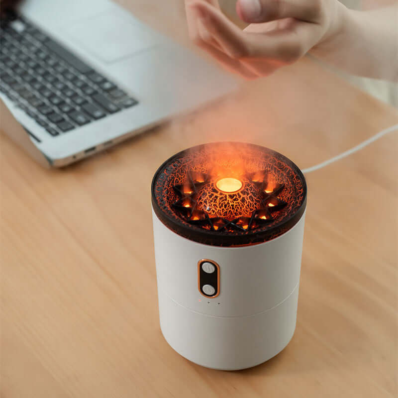 Aroma diffuser with LED flame effect on a desk near a laptop, releasing mist.