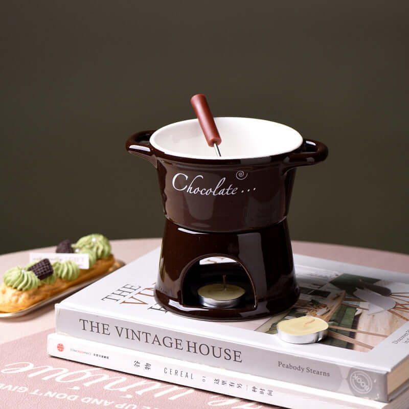 Ceramic chocolate fondue pot with fork and tealight on books, dessert in background