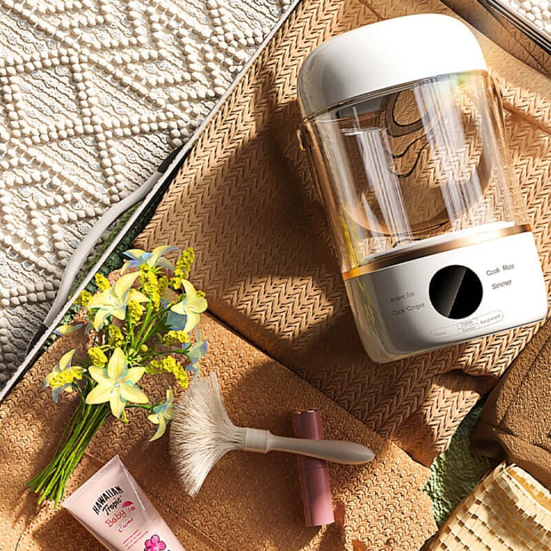 Compact electric rice cooker on woven mat with artificial flowers and kitchen tools