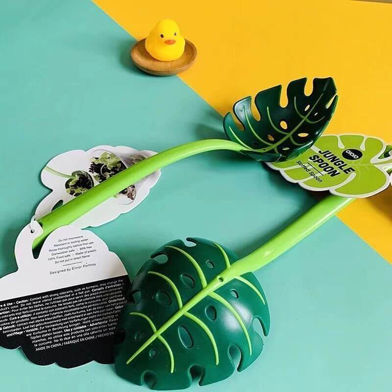 Leaf-shaped kitchen serving spoon with green handle on colorful background, rubber duck in dish nearby