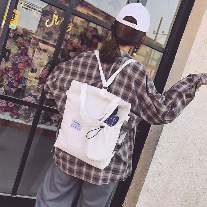 Woman in plaid shirt and white cap wearing a white canvas backpack outside a flower shop
