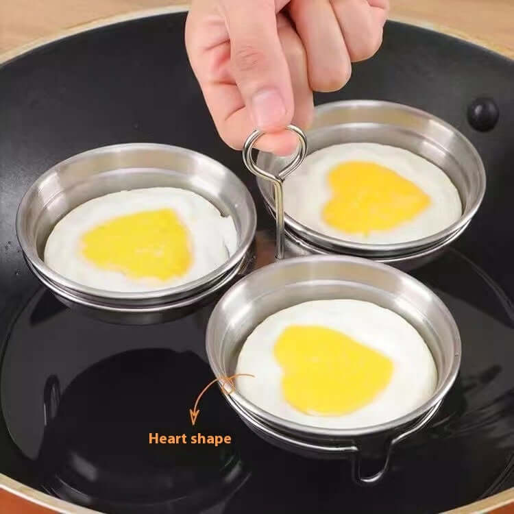 Stainless steel egg rings making heart-shaped fried eggs in a nonstick frying pan.