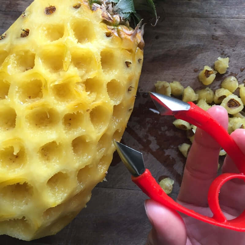 Hand using a pineapple eye remover tool to core pineapple eyes on a wooden cutting board.