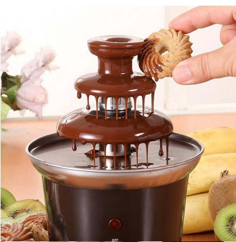 Mini chocolate fountain with cookie being dipped, surrounded by fruits in kitchen setting