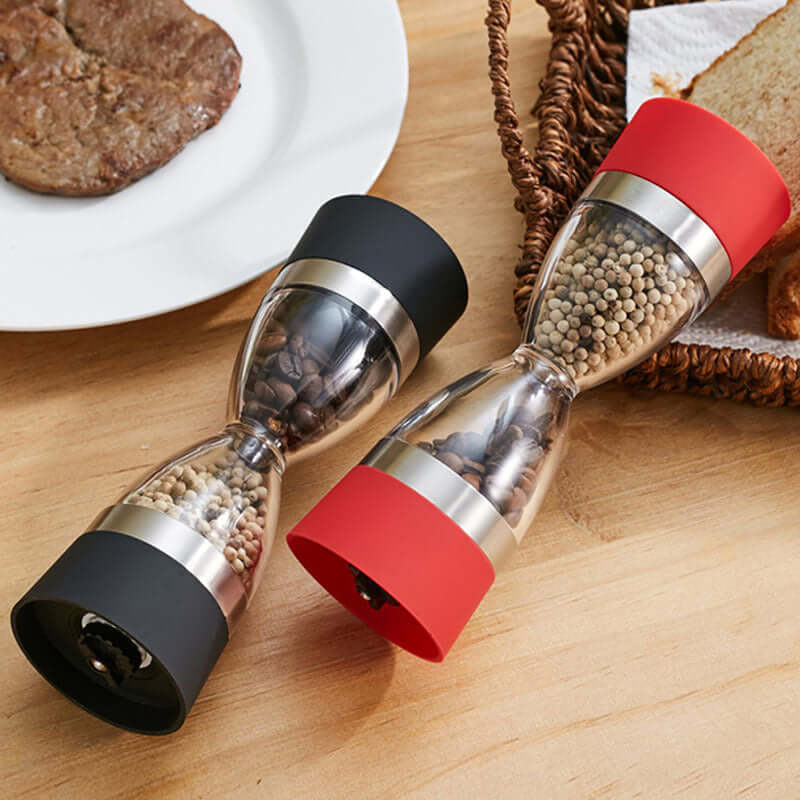 Dual manual spice grinders with black and red ends on wood table, filled with peppercorns and coffee beans