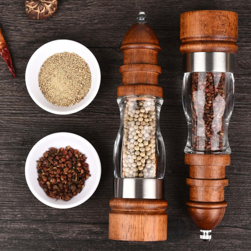 Wooden spice grinders with peppercorns and small bowls of ground spices on a dark kitchen counter
