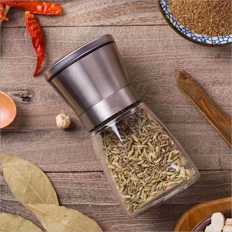 Stainless steel spice grinder filled with fennel seeds on a wooden kitchen counter with herbs and spices.