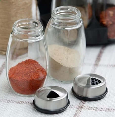 Glass spice jars with metal shaker lids, filled with red and white seasoning, on kitchen counter