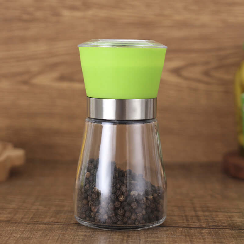 Glass pepper grinder with green lid on wooden kitchen countertop, filled with black peppercorns.