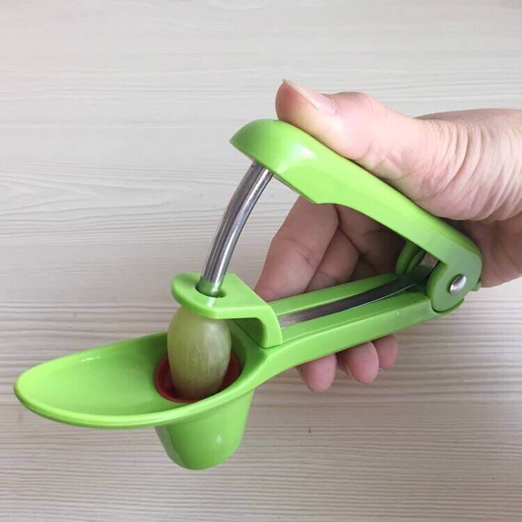 Hand using green kitchen tool to pit an olive or cherry on light wood surface