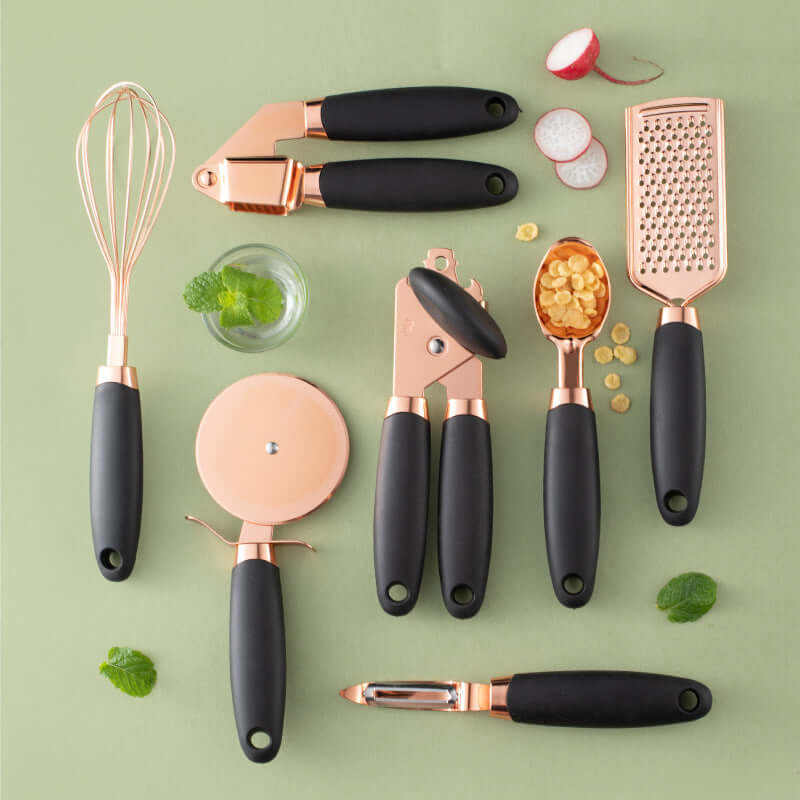 Rose gold and black kitchen utensil set with whisk, garlic press, grater, peeler, can opener, pizza cutter, and ice cream scoop on a green background
