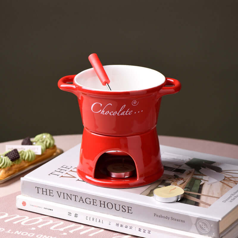 Red ceramic chocolate fondue pot with fork on books, tabletop kitchen accessory