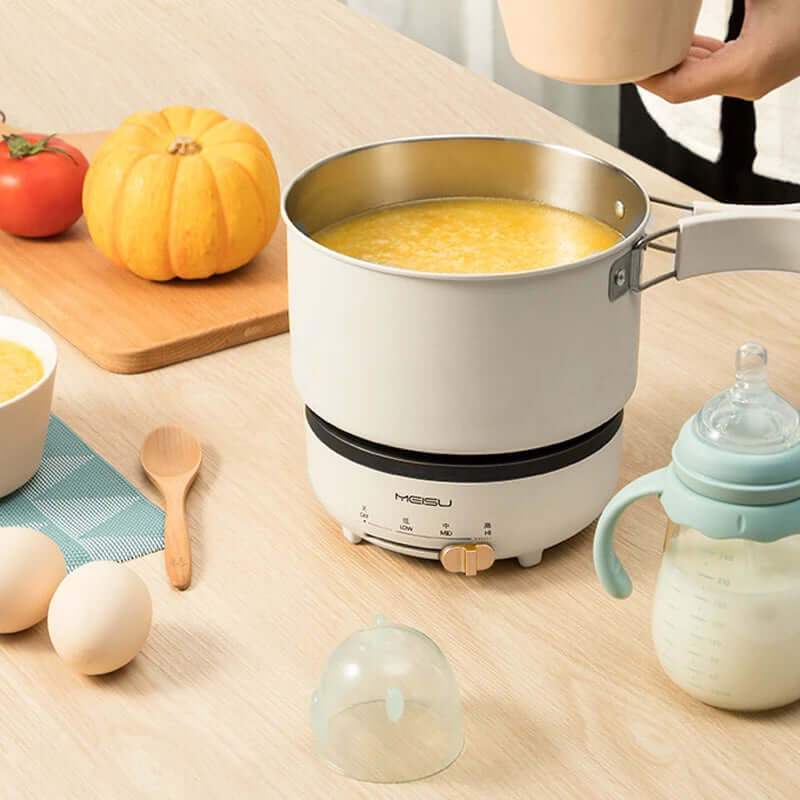Electric cooking pot with pumpkin soup on a kitchen table, surrounded by eggs, baby bottle, and vegetables