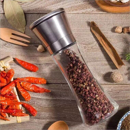 Stainless steel spice grinder filled with peppercorns on a wooden kitchen table with dried chili peppers and herbs.