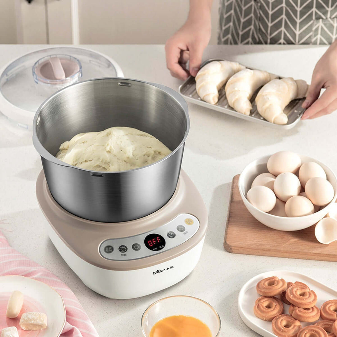 Electric dough mixer with stainless steel bowl on kitchen counter, surrounded by eggs, croissants, and pastries.