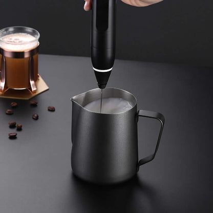 Handheld milk frother foaming milk in stainless steel pitcher with coffee in background