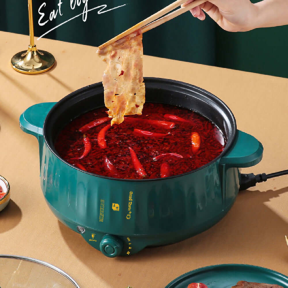 Electric hot pot with spicy broth and chili peppers, cooking meat using chopsticks.